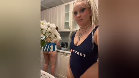 Media: Video of a white woman with long blonde dreadlocks wearing a blue \"NastyBET\" swimsuit, standing in a kitchen, holding a bouquet of white flowers.