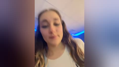 Media: A blurry video of a young woman with light skin and long brown hair, wearing a white shirt, indoors with a blue light background.