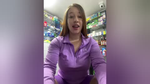 Media: Video of a young, light-skinned woman with shoulder-length brown hair, smiling in a grocery store aisle. She wears a lavender zip-up jacket, standing in front of colorful, stacked shelves filled with various food products.