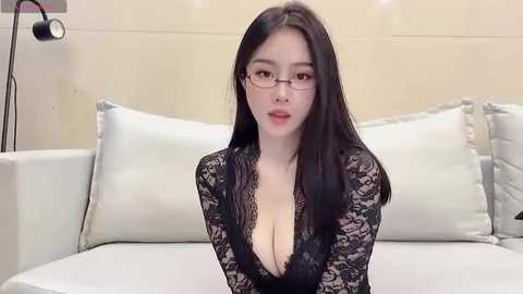 Media: Video of an Asian woman with long black hair, fair skin, wearing glasses, black lace dress, sitting on a white sofa in a modern, minimalist room.