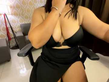 Media: Video of a curvy, light-skinned woman with black hair, wearing a black bra and matching skirt, seated in a gray chair in a room with patterned wallpaper.