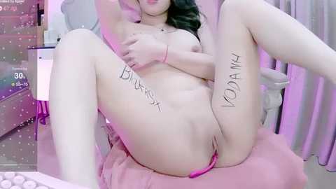 Media: A video of a nude Asian woman with medium-sized breasts, lying on her back with legs spread, displaying her shaved vulva. She has \"BUNNY SEX\" tattooed on her thighs. The background is a dimly lit room with a pink sheet.