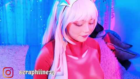 Media: Video of a woman in a red latex bodysuit and cat ears, posing against a blue and pink background, with the username \"seraphine23\" in the corner.