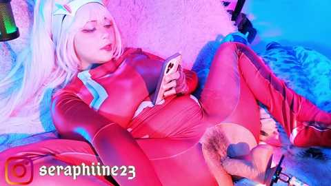 Media: Video of a fair-skinned woman with long platinum blonde hair, dressed in a shiny red latex bodysuit, sitting on a bed, holding a phone, with blue and pink lighting, and the username \"seraphine23\" in the corner.