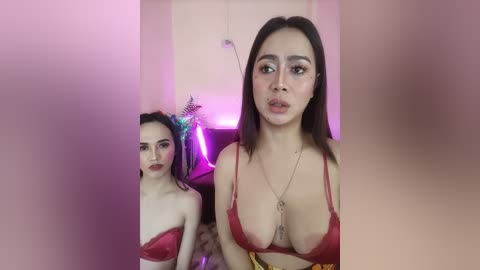 Media: Video of two young women with fair skin, one wearing a red bra, the other in a blue bra, standing indoors against a blurred background.