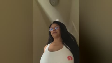 Media: Video of a curvy woman with long black hair, wearing glasses, a white tank top, and a red logo on her chest. She stands in a beige hallway with a wall clock and dim lighting.