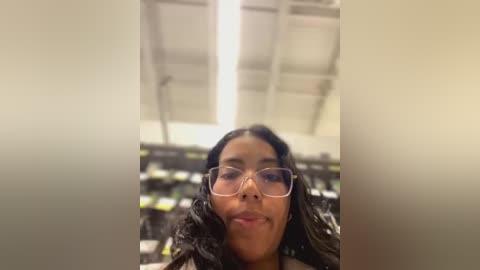 Media: A video of a young woman with long, dark hair, wearing glasses, and a smile, standing in a dimly lit room with a ceiling light and blurred bookshelves in the background.