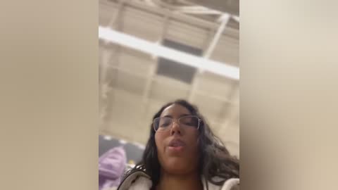 Media: Video of a young woman with long black hair, wearing glasses, and a white jacket, leaning against a beige wall in an indoor setting with fluorescent lighting.