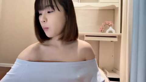 Media: A video of an East Asian woman with straight black hair, wearing an off-shoulder white top, in a simple, beige-colored room with a white shelf and a pink flower decoration.