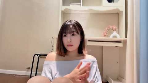 Media: Video of an Asian woman with shoulder-length brown hair, wearing a white off-shoulder top, sitting on a stool in a beige room with a desk and floral decor.