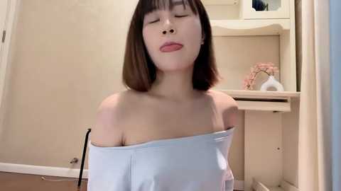 Media: A video of an Asian woman with straight, shoulder-length brown hair, wearing a white off-shoulder top and a nose ring, in a bathroom with beige walls and a white sink.