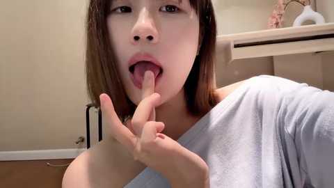 Media: A video of an East Asian woman with straight brown hair, light skin, and an off-the-shoulder light gray shirt, licking her middle finger in a provocative manner against a beige wall and wooden vanity.