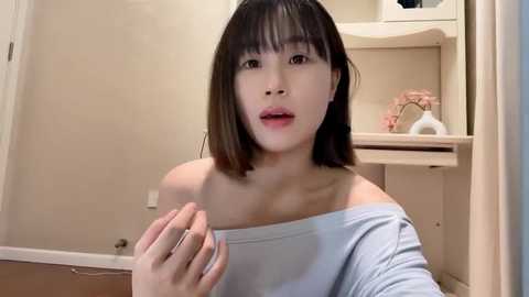 Media: Video of a young Asian woman with straight, shoulder-length black hair, wearing a light blue off-shoulder top. She is indoors, possibly in a bathroom, with beige walls and wooden cabinets in the background.