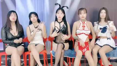 Media: Video of five Asian women in revealing lingerie and bunny costumes, sitting on red stools against a white backdrop, with a modern stage setup.