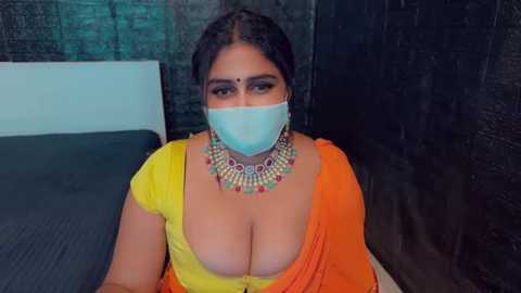 Media: Video of a South Asian woman with medium skin tone, wearing an orange sari with a plunging neckline, large earrings, and a face mask, sitting on a bed with a dark wall and gray sheets.