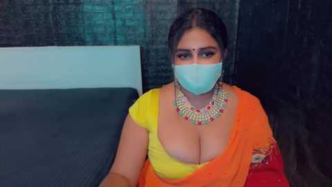 Media: A video shows a South Asian woman in a yellow and orange sari, wearing a face mask, large earrings, and a decorative necklace. She sits on a bed in a dimly lit room with dark walls and a green curtain.