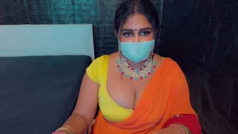 Media: Video of a South Asian woman with medium brown skin, wearing a yellow and orange sari, gold jewelry, and a face mask, seated on a dark bed with a textured brick wall background.