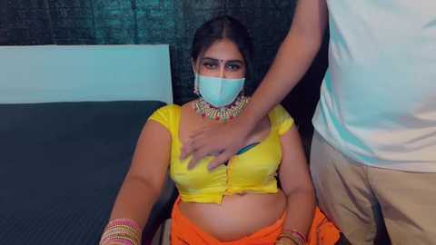 Media: Video of a South Asian woman with medium skin tone, wearing a yellow crop top, orange skirt, and face mask, being held by a man in a white shirt and beige pants.