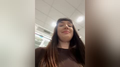 Media: A video of a young woman with long, straight, dark brown hair and fair skin, wearing a brown top, smiling in a pharmacy with white ceiling tiles and fluorescent lights.