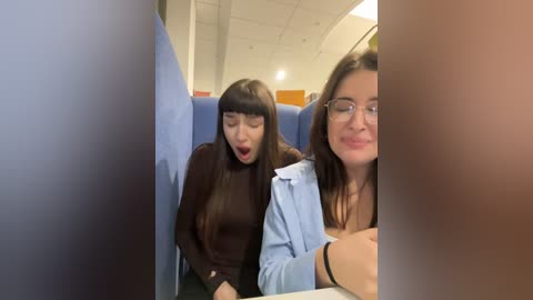 Media: Video of two young women, one with long dark hair, wearing glasses, and the other with straight hair, sitting in blue airline seats, smiling and laughing.