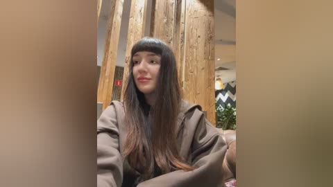 Media: A video of a young woman with straight, dark brown hair and blunt bangs, wearing a brown coat, seated in a wooden interior with exposed beams.