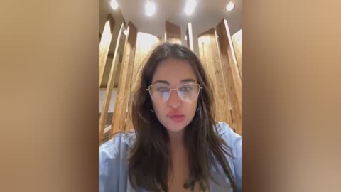 Media: Video of a young woman with long brown hair, wearing glasses, light blue shirt, standing in a wooden structure with tall, cylindrical walls.