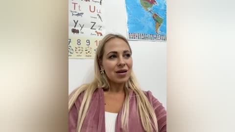 Media: Video of a smiling, blonde woman with long hair, wearing a pink cardigan and white top, standing in a room with educational posters.