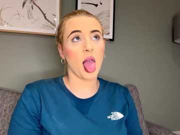 Video of a blonde woman with fair skin, wearing a blue The North Face sweatshirt, sticking out her tongue in a playful manner. Background features gray couch and framed art on light green wall.