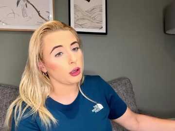 Media: Video of a blonde woman with fair skin, wearing a blue t-shirt, and a silver necklace, seated on a gray couch. The background features minimalist art on a muted green wall.