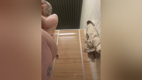 Media: Video of a nude blonde woman with a tattoo on her thigh, standing in a narrow hallway with beige and wooden walls. She has her back to the camera, facing a beige coat draped over a chair.