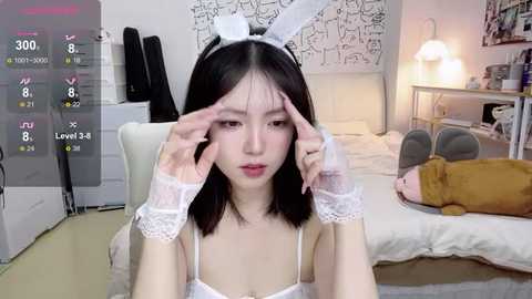 Media: Video of an East Asian woman with straight black hair, wearing a white lace bunny headband and matching lace cuffs, adjusting her hair in a bedroom with white walls, a bed, and a lamp.