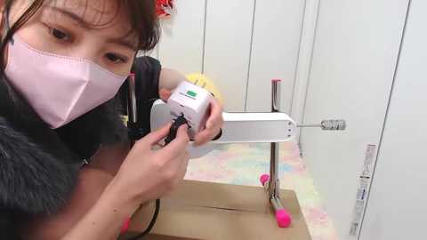 Media: Video of an Asian woman in a pink surgical mask, black jacket, and green wristband, adjusting a white medical device on a pink stand in a sterile, white-tiled room with a colorful floor.