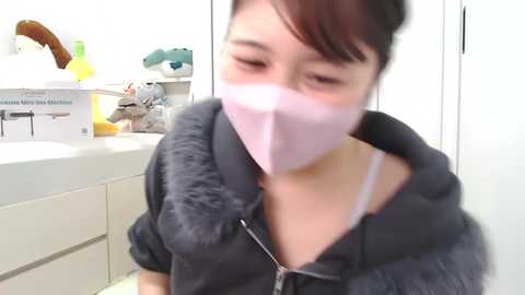 Media: A video of an Asian woman with short black hair, wearing a pink surgical mask and a black fur-lined hoodie, standing in a white bathroom with stuffed animals on a counter.