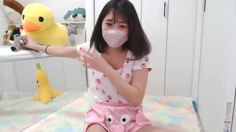 Media: Video of an East Asian woman with shoulder-length black hair, wearing a pink face mask, white shirt, and pink shorts with cartoon eyes, sitting on a pastel-colored blanket in a room with white walls, plush toys, and a yellow duck.