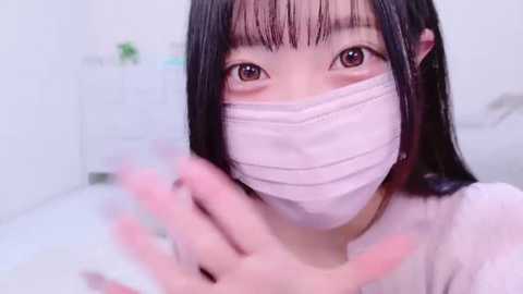 Media: Video of an East Asian woman with long black hair, wearing a white mask, touching her face with a hand, in a white room.