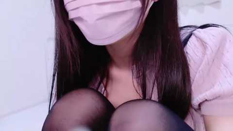 Media: Video of an Asian woman with long, straight dark hair, wearing a pink surgical mask and a light pink blouse. She is seated, with her legs crossed, and her breasts partially covered by sheer black stockings. The background is a plain, light-colored wall.