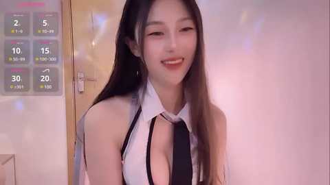 Media: Video of an East Asian woman with long, straight black hair, wearing a revealing white top with a black tie, smiling in a dimly lit room with a calendar displaying weather and temperature.