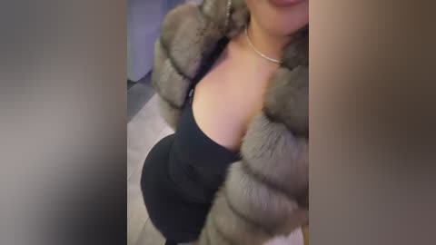 Media: Video of a woman with light skin, wearing a black top and a fur-lined coat, standing in a dimly lit room.