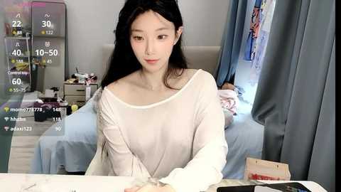 Media: A video of an East Asian woman with long black hair, wearing a sheer white top, sitting at a desk, with a baby in a crib in the background.