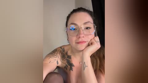 Media: Video of a fair-skinned woman with glasses, brunette hair, tattoos on her shoulders, and wearing sheer black stockings, leaning against a black curtain in a dimly lit room.