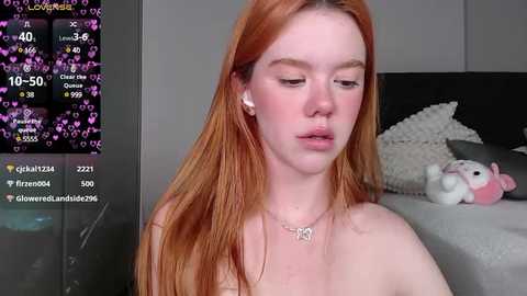 Media: Video of a pale-skinned, red-haired woman with freckles, wearing white earbuds, sitting on a bed with a stuffed animal, in a dimly lit room.