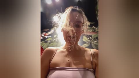 Media: Video of a young woman with fair skin, messy blonde hair, wearing a light purple spaghetti strap top. Background features blurred, dark indoor setting with dim lighting and plant leaves.