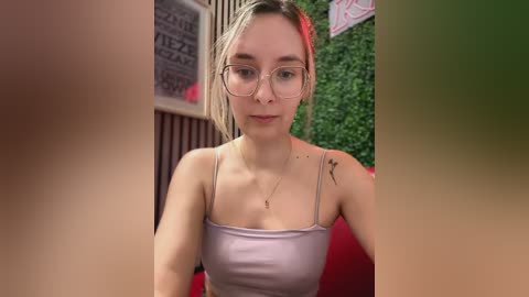 Media: Video of a young Caucasian woman with light skin and blonde hair, wearing glasses and a light pink spaghetti strap top, sitting indoors with greenery and red seating in the background.