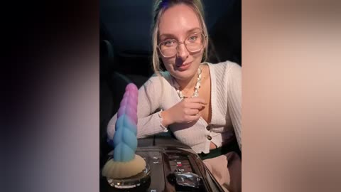 Media: Video of a blonde woman with glasses, wearing a white cardigan and necklace, holding a blue and pink dildo in her car, with dim lighting.