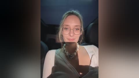 Media: Video of a woman with light skin, blonde hair, and glasses, wearing a white top and a dark necklace, sitting in a car with dim lighting.