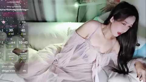 Media: Video of a young Asian woman with long black hair, wearing a light pink, off-shoulder dress, lying on a bed, with virtual reality glasses overlaying the scene.