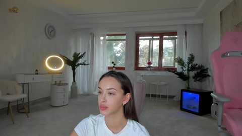 Media: A video of a young woman with fair skin and straight brown hair, sitting in a modern, minimalist bedroom with white walls, large windows, and a pink gaming chair.