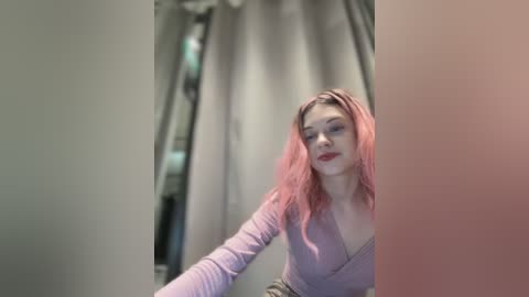 Media: Video of a young woman with fair skin and long, wavy pink hair, wearing a light purple V-neck top, standing indoors with grey curtains in the background.