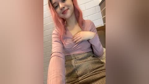 Media: Video of a young woman with pink hair, wearing a pink long-sleeve top and a checkered skirt, smiling, standing in a tiled bathroom.