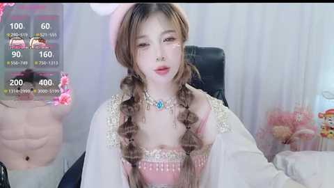 Media: Video of an East Asian woman with fair skin, long brown hair in braids, wearing a pink corset, white jacket, and a necklace, sitting on a black chair, in a soft, pastel-toned room.